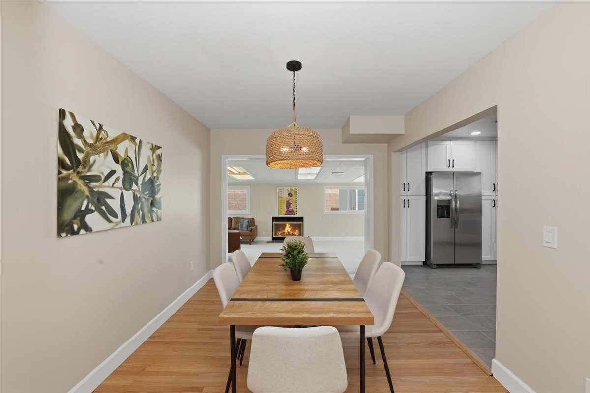 Staged dining room with a wood table, white upholstered chairs and a woven pendant light, opening to a family room with a fireplace
