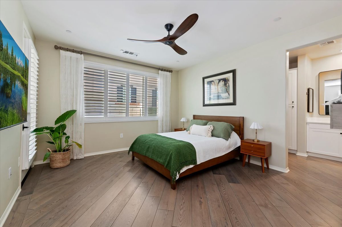 Staged primary bedroom with a wood bed and green throw, plantation shutters, ceiling fan, wide-plank wood floors and a view into the bathroom